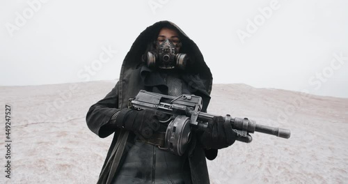 Slow motion post apocalyptic woman in black hooded mantle and grunge protective gas mask posing with gun standing on hill in white dead desert. Cyberpunk future warrior female survivor after war photo