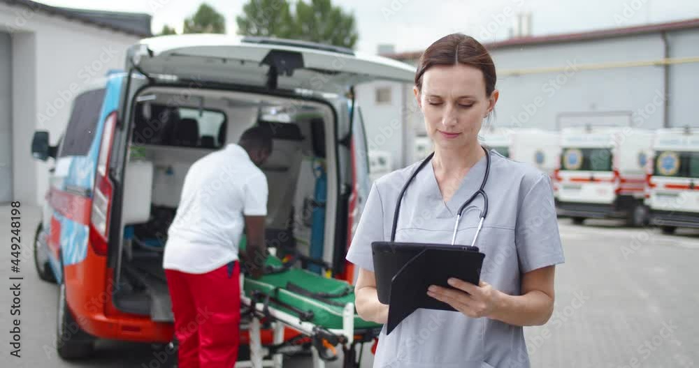 Female doctor filling in medical form and using table device. Outdoor. African American doc working at ambulance on background. Woman medic tapping and scrolling on computer online. Team of medics. Female doctor filling in medical form and using table device. Outdoor. African American doc working at ambulance on background. Woman medic tapping and scrolling on computer online. Team of medics.