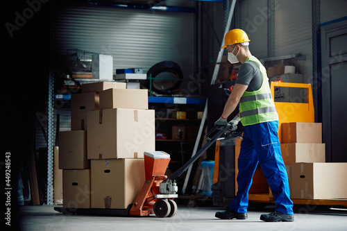 Full length of worker pushes pallet jack while working warehouse during coronavirus pandemic.