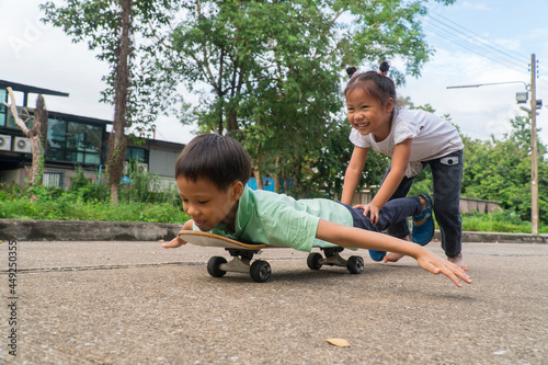 Small asain teenage girl playing with brother pushing forward boy riding on skateboard in public road. Kids and entertainment. Chilhood and sports concept.
