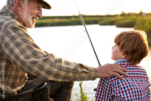 Portrait of awesome handsome fgrandather with grandson sitting on lake while enjoying fishing together and laughing in rays of sunset sunlight, in the evening. copy space for text