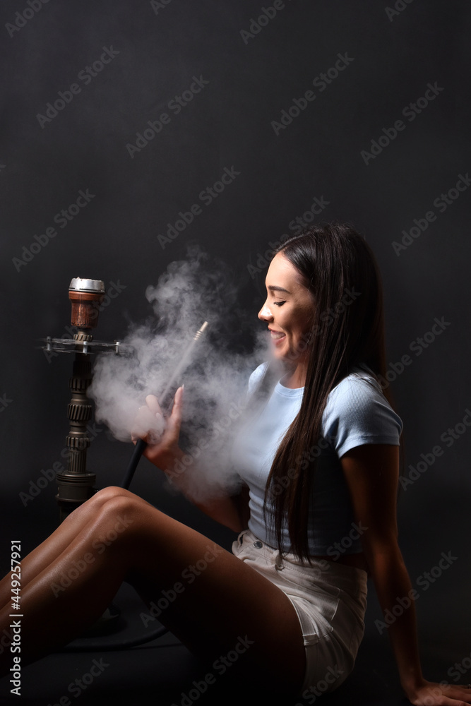 Obraz premium The girl smokes a hookah on a black background. Smoke comes out of the mouth. The pleasure of smoking 