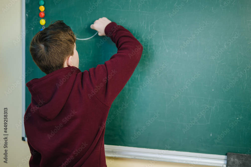 Student of higher school is standing in dark hoodie near the green blackboard in the classroom writing alphabet . View from the back. School education concept Student of higher school is standing in dark hoodie near the green blackboard in the classroom writing alphabet . View from the back. School education concept