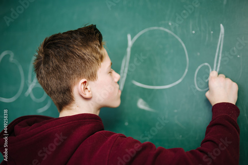 Student of higer school is standing in dark hoodie near the green blackboard in the classroom. Teenage boy is writing alphabet on the board. School education concept photo