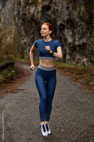 Running outdoors. Redhead Runner woman jogging in spring forest. Beautiful young fit fitness sport model jogging in sportive outfit. Attractive caucasian lady engaged in sport workout, on fresh air