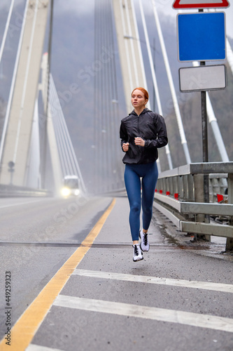Young redhead confident female in sportive outfit jogging in urban area at spring day on bridge, looking at side while running, focused on sport. side view full-length photo, copy space