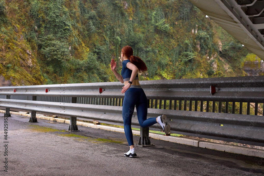 Rear view On Fit redhead Powerful lady jogging on modern bridge, view from back. urban sports, running in the morning, outdoors. copy space. sport, healthy lifestyle. sport and activity concept