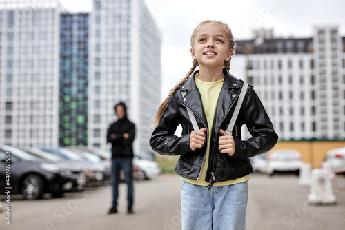 Positive child girl walking alone on street, while terrible maniac looking at her in the background. Focus on kid girl in casual leather jacket enjoy walk, don't afraid of adult men. kidnapping photo
