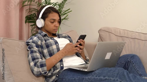 A young woman in jeans using a silver laptop and smartphone while lying on the couch. The concept of working and learning online.