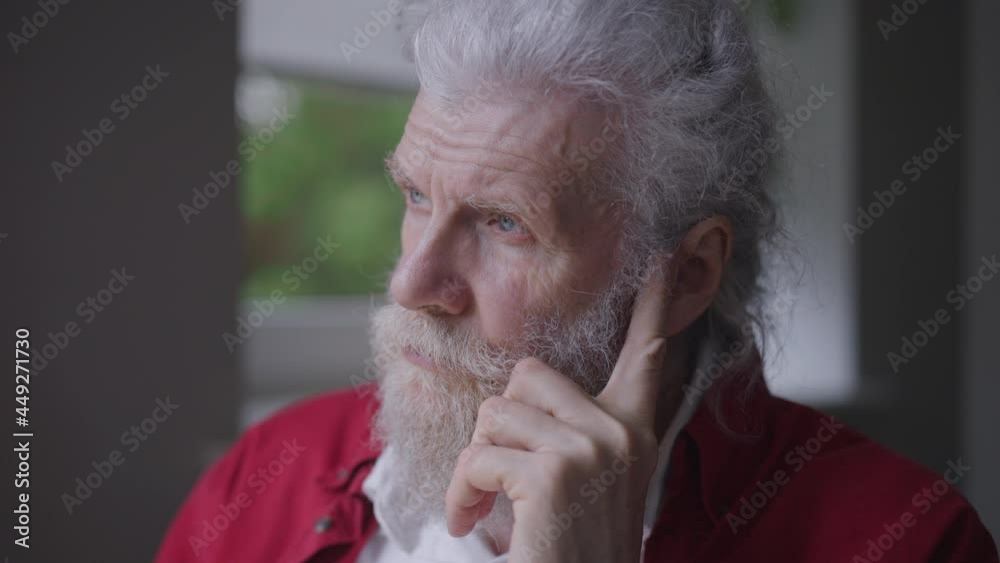 Close-up portrait of handsome old Caucasian bearded grey-haired man with blue eyes looking out the window at home holding head in hands. Headshot of depressed sad lonely retiree alone indoors Close-up portrait of handsome old Caucasian bearded grey-haired man with blue eyes looking out the window at home holding head in hands. Headshot of depressed sad lonely retiree alone indoors