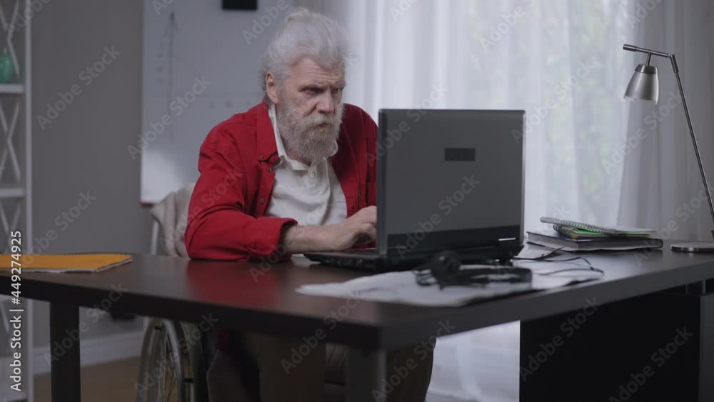 Serious concentrated disabled man in wheelchair typing on laptop keyboard messaging online from home office. Portrait of professional focused Caucasian invalid on remote working indoors. Inclusivity Serious concentrated disabled man in wheelchair typing on laptop keyboard messaging online from home office. Portrait of professional focused Caucasian invalid on remote working indoors. Inclusivity