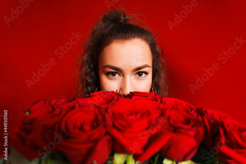 Portrait of girl peeking out from behind bouquet of flowers on red background. Portrait of girl peeks out from behind bouquet of flowers on red background. Young woman holding large bouquet red roses photo