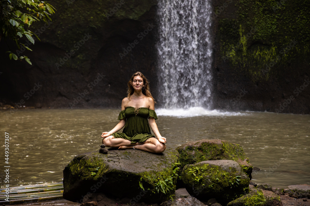 Yoga lotus pose. Young Caucasian woman sitting on the stone, meditating, practicing yoga, pranayama at waterfall. Hands in gyan mudra. Closed eyes. Yoga retreat. Tibumana waterfall, Bali