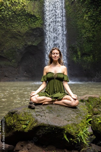 Yoga lotus pose. Young Caucasian woman sitting on the stone  meditating  practicing yoga  pranayama at waterfall. Hands in gyan mudra. Closed eyes. Yoga retreat. Tibumana waterfall  Bali
