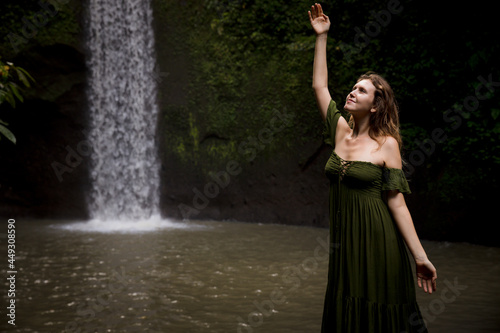Portrait of Caucasian woman near the waterfall. Hand raised up. Enjoy nature. Water splash. Woman wearing green dress. Travel to Asia. Copy space. Tibumana waterfall  Bali