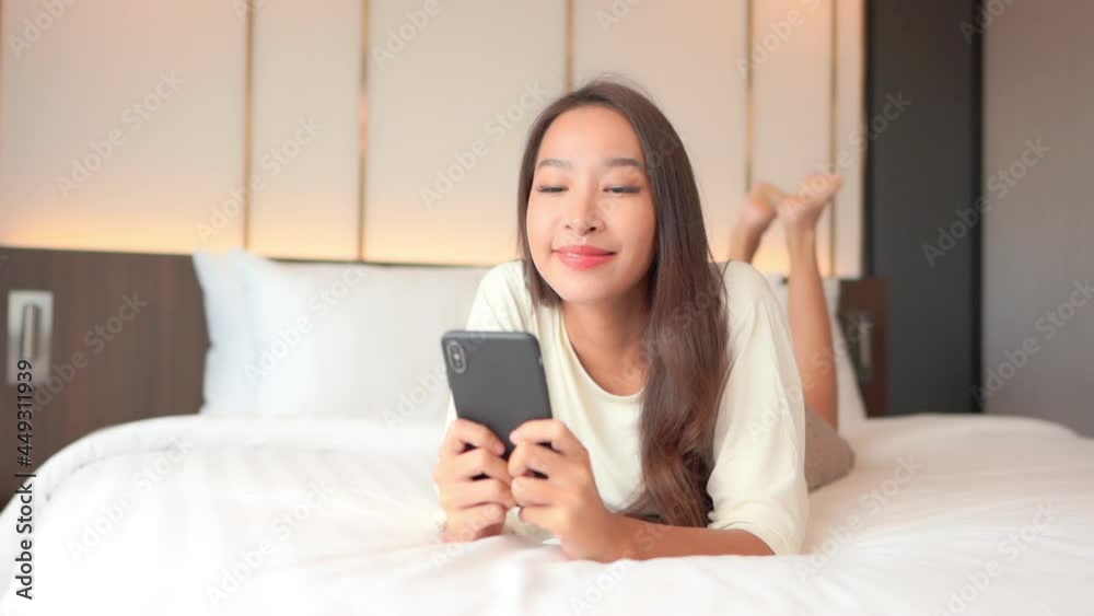 Face Close-up on Asian woman watching video on mobile phone holding smartphone with both hands while comfortably lying on a bed in a hotel room Face Close-up on Asian woman watching video on mobile phone holding smartphone with both hands while comfortably lying on a bed in a hotel room