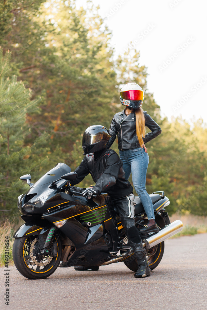 Motorcyclist in a leather jacket sits on a motorcycle and a girl stands on a motorcycle in helmets against the background of trees in the forest
