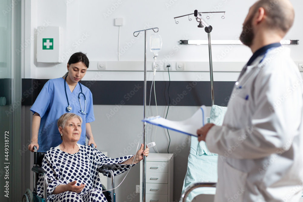 Obraz premium Sick senior woman hospitalized sitting in wheel chair with iv drip bag attached on arm, helped by nurse to move. Doctor with stethoscope checking patient before surgery and talking about treatment