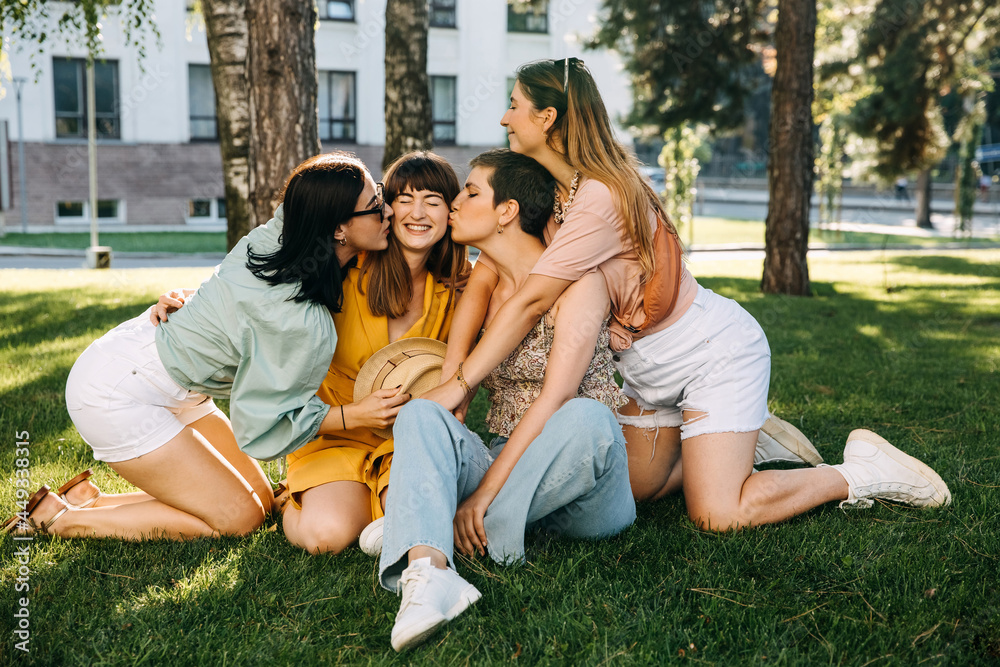 Group of four friends sitting in a park on green grass, hugging and smiling.
