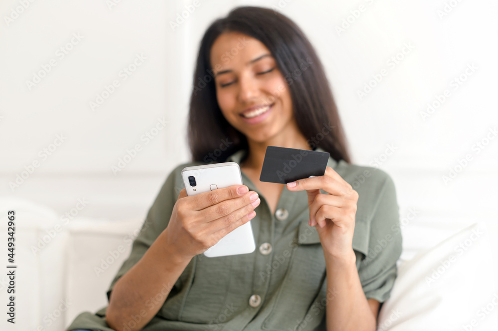 Cheerful young indian woman using smartphone shopping app for buying online, holding credit card and inputs banking data, sits on sofa in relaxed atmosphere in living room at home, headshot Cheerful young indian woman using smartphone shopping app for buying online, holding credit card and inputs banking data, sits on sofa in relaxed atmosphere in living room at home, headshot