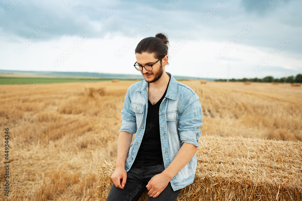 Confident smiling guy in denim jacket sitting on haystacks in farm field.