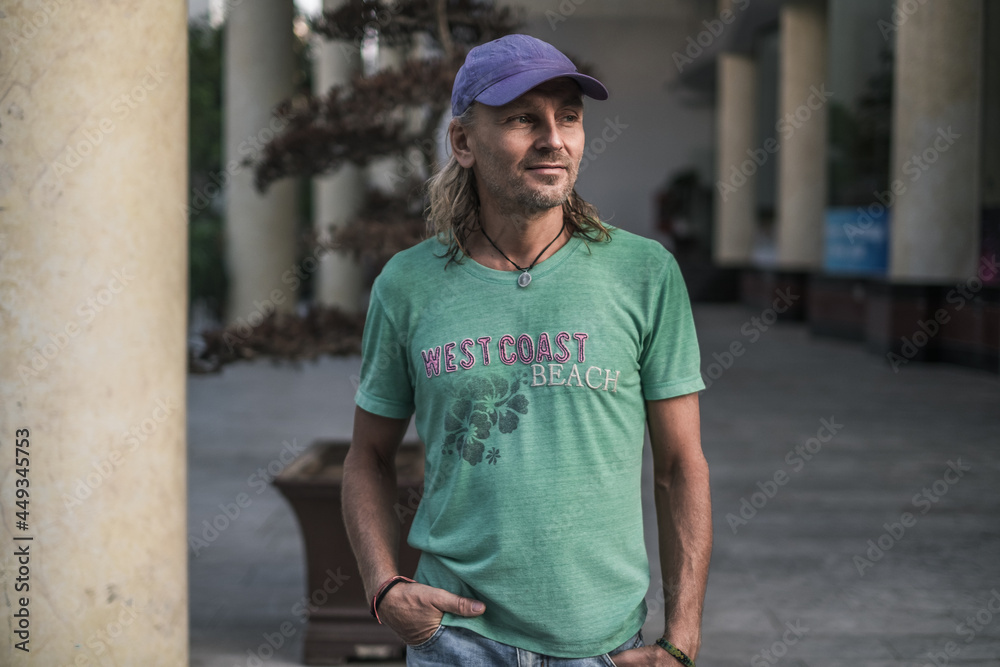 Mature man with long grey hair wearing green t-shirt and blue cap standing on the street in town and looking aside. Portrait. Mature man with long grey hair wearing green t-shirt and blue cap standing on the street in town and looking aside. Portrait.