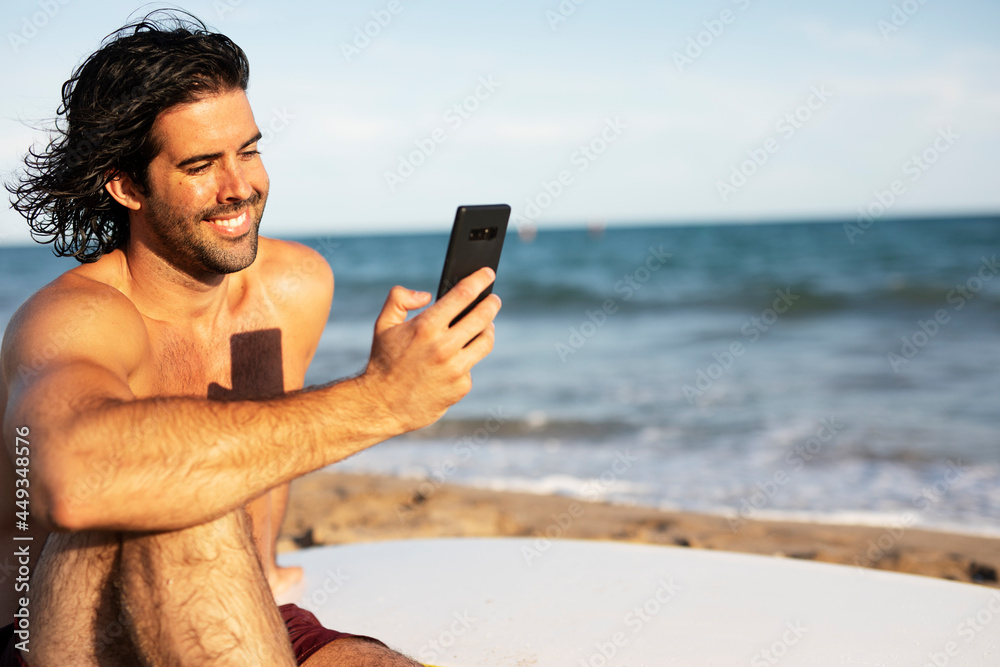 Portrait of handsome surfer with his surfboard. Young man using the phone while relaxing on the beach