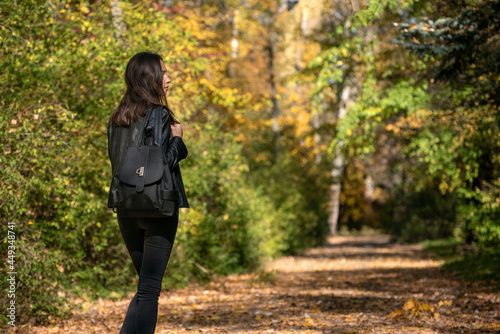 Brunette girl with a backpack walks through the autumn forest. Young woman rests in the park. Back view