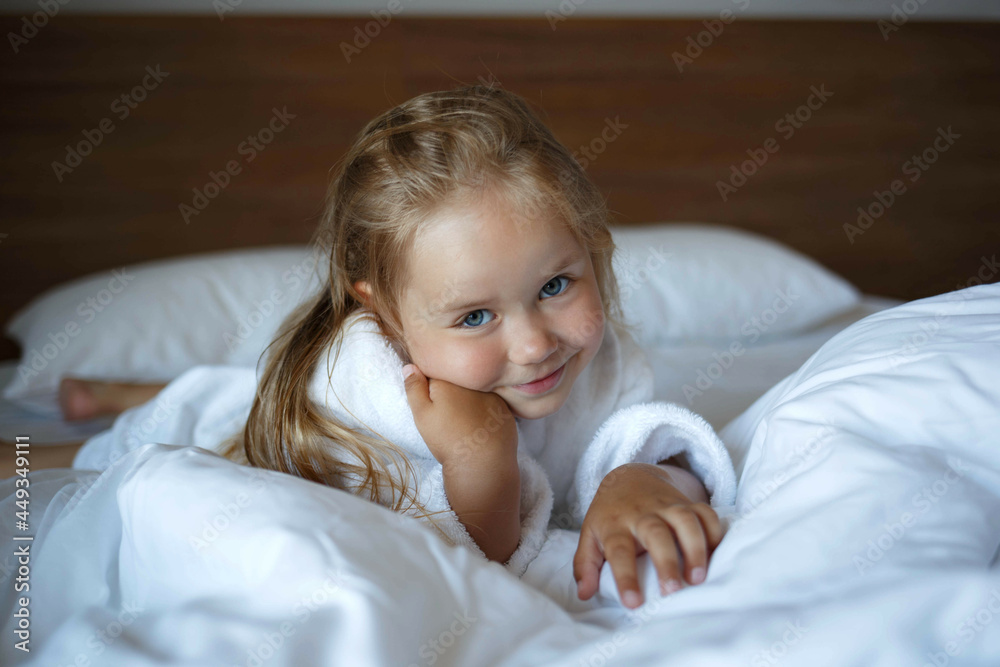 portrait of a little cute girl in a white robe lying on the bed in the bedroom portrait of a little cute girl in a white robe lying on the bed in the bedroom