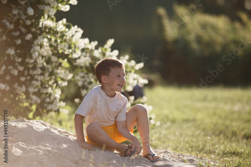 a boy in yellow shorts and a light T-shirt is sitting on the sand near a bush with white flowers, laughing, and the sun is shining on him