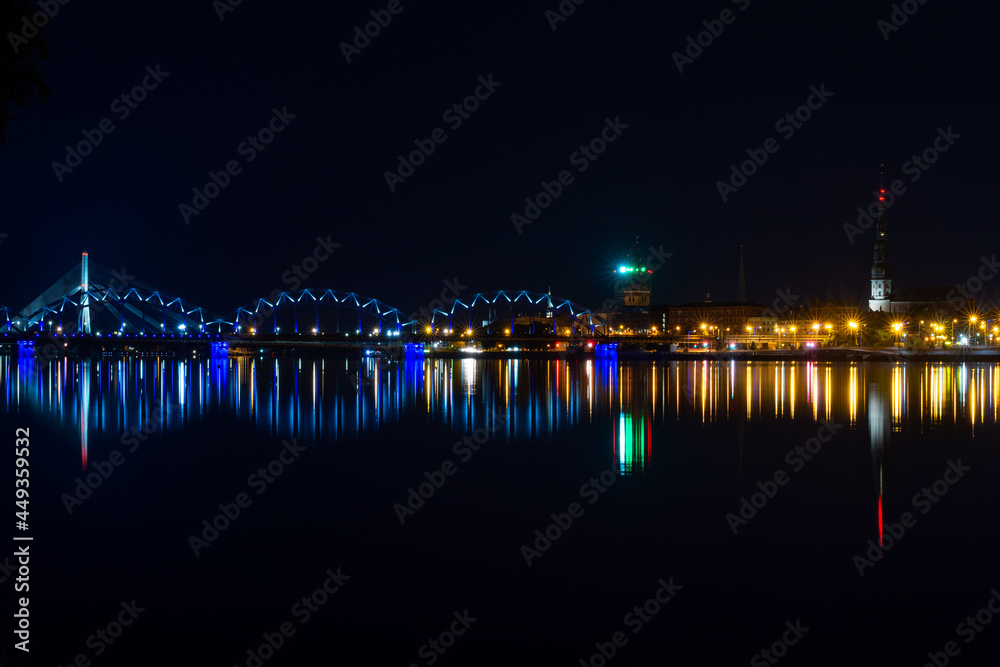View of Riga (Latvia) in the evening. Old buildings and a railway bridge are illuminated and reflect in the Daugava river.