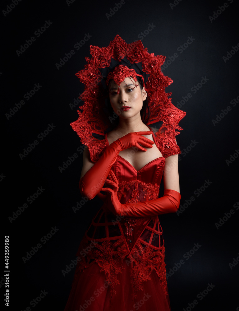Full length  portrait of beautiful young asian woman wearing red corset, long opera gloves and ornate gothic queen crown. Graceful posing isolated on a dark studio background.