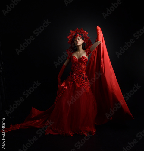 Full length portrait of beautiful young asian woman wearing red corset, long opera gloves and ornate gothic queen crown. Graceful posing isolated on a dark studio background.