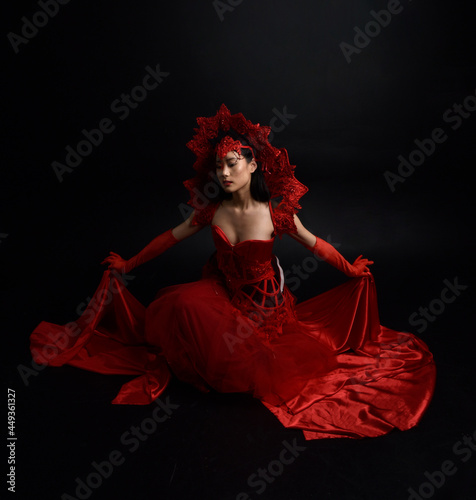 Full length portrait of beautiful young asian woman wearing red corset, long opera gloves and ornate gothic queen crown. Graceful posing isolated on a dark studio background.