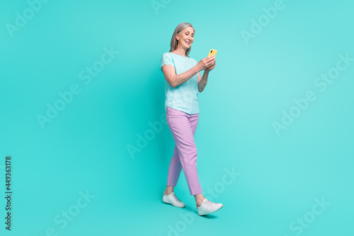Full size profile side photo of smiling mature woman go walk addicted to smartphone isolated on turquoise color background