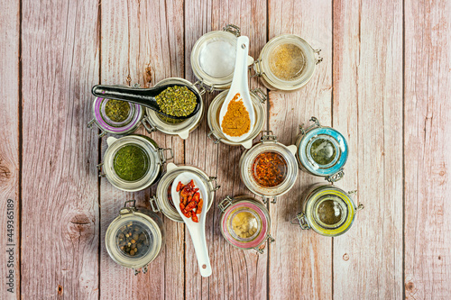 Transparent glass jars with assorted spices seen from the top with some spoons with yellow curry, small cayenne and oregano. Sea salt flakes, white pepper, sesame seeds
