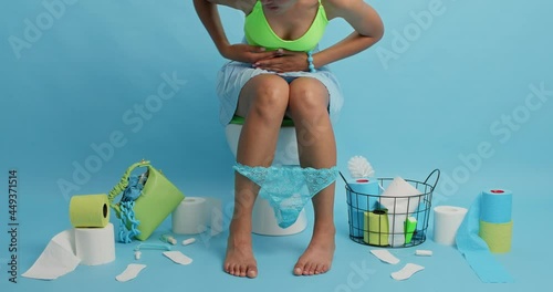 Unknown woman suffers from abdominal cramps takes pills has severe stomachache poses on toilet bowl surrounded by hygiene products isolated over blue wall feels discomfort. Food poisoning gastritis