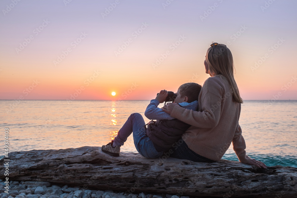 Mother and son tourist traveling sitting on the beach and look binocular under sunset sky with sun in summer. Mothers Day.