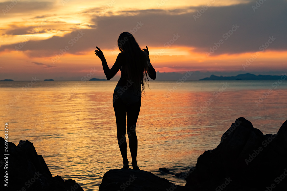 Silhouette of a young woman dancing on the rocks by the sea at colorful sunset background