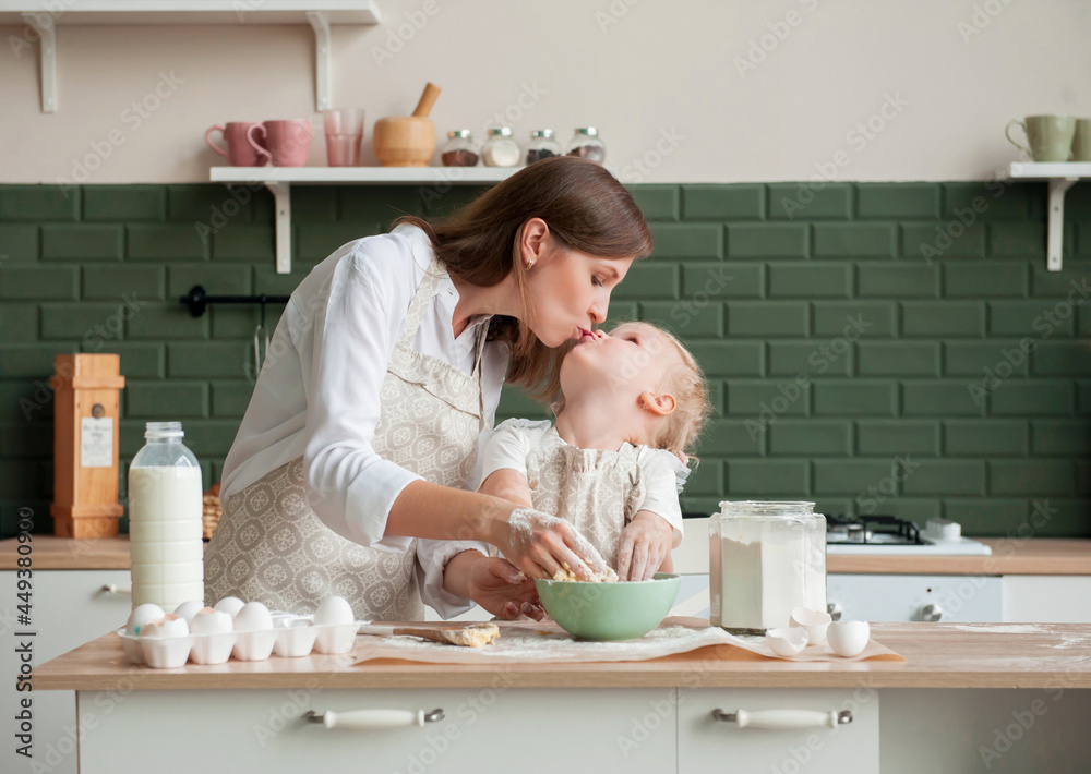 Happy mother and her little daughter knead dough together at home in the kitchen. Little girl kisses mom and helps her to cook