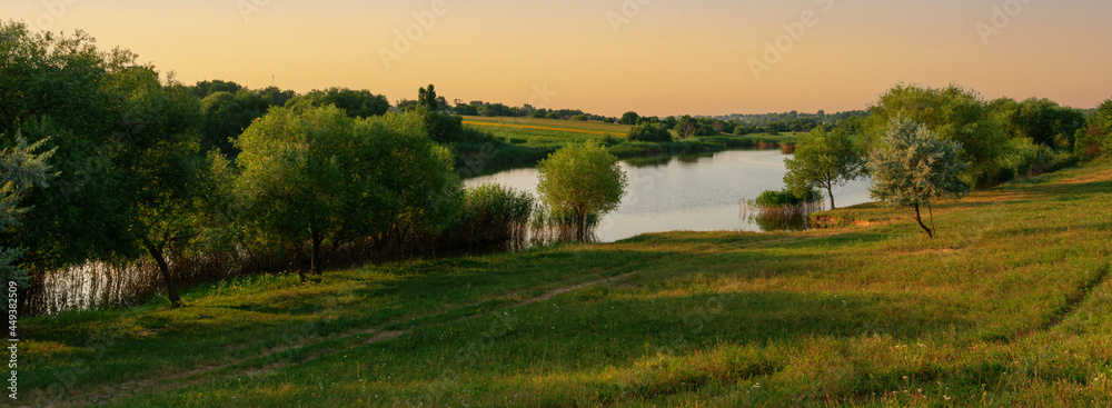 Rural landscape, panorama, banner - small country lake with a meadow and green forest in the background Rural landscape, panorama, banner - small country lake with a meadow and green forest in the background