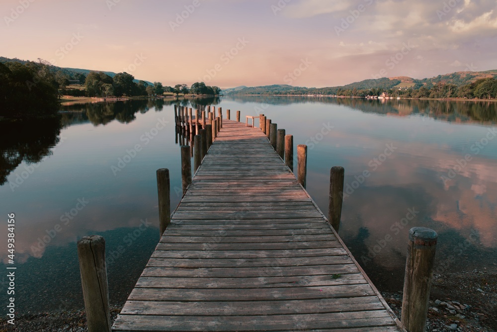 Soft pink morning light falls washes over a wooden jetty on Coniston Water in the Lake District, reflecting clouds on the surface of the lake