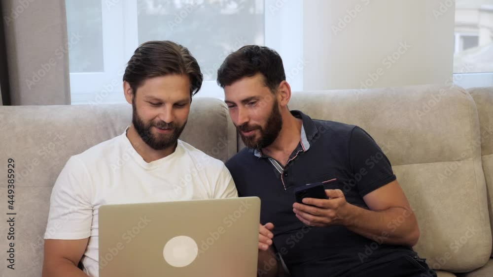 Young same sex loving couple sitting on the sofa at home using mobile phone and laptop for social media online shopping. Homosexual men spending time together relax using technology play games.