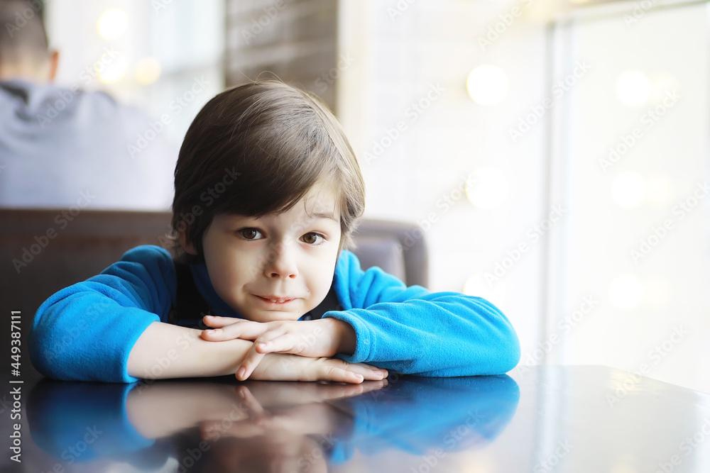 A young boy sits at a table near the window in a cafe and waits for a waiter to come up and take an order. A young boy sits at a table near the window in a cafe and waits for a waiter to come up and take an order.