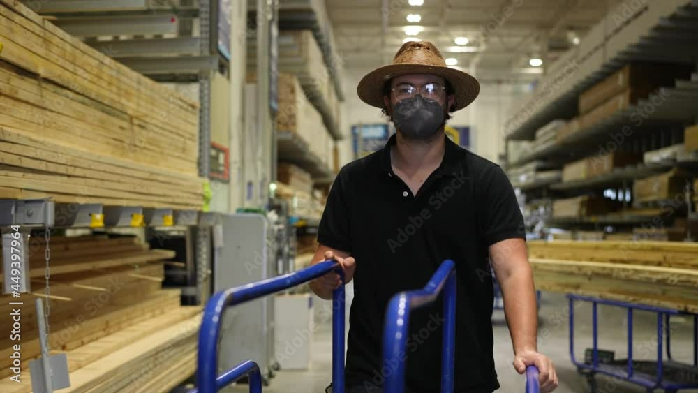 Wearing a face mask, a man pushes a lumber cart down an aisle full of wood products at a big box hardware store.