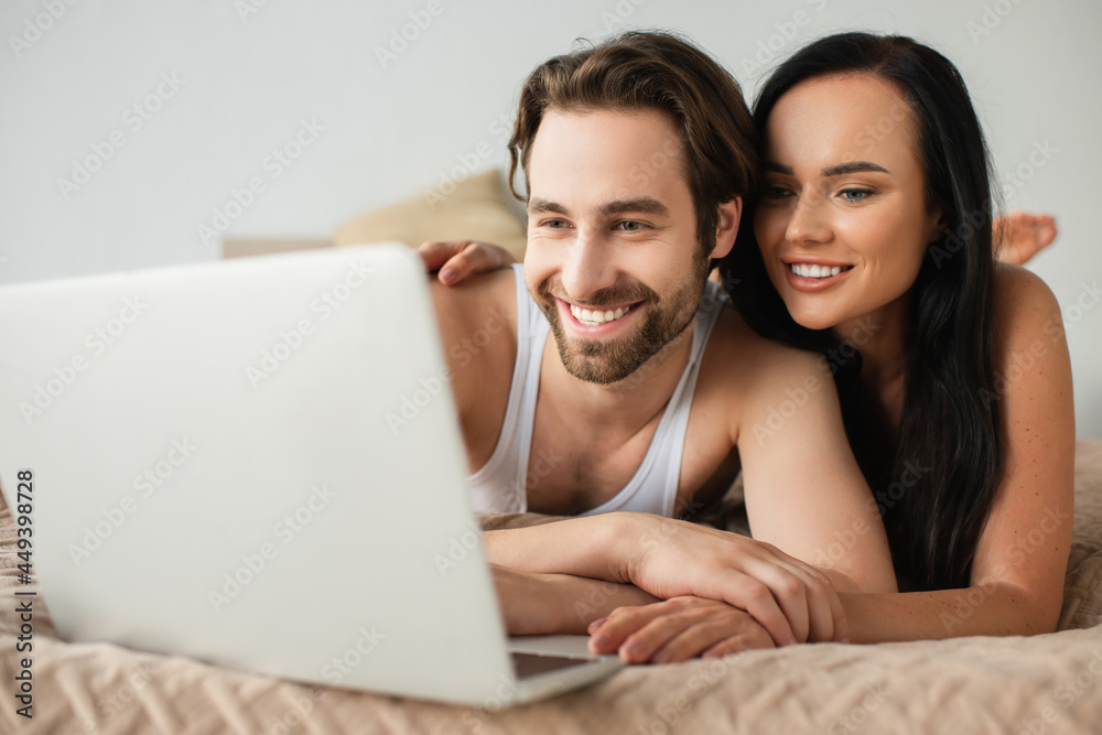joyful couple watching movie on blurred laptop while lying on bed joyful couple watching movie on blurred laptop while lying on bed