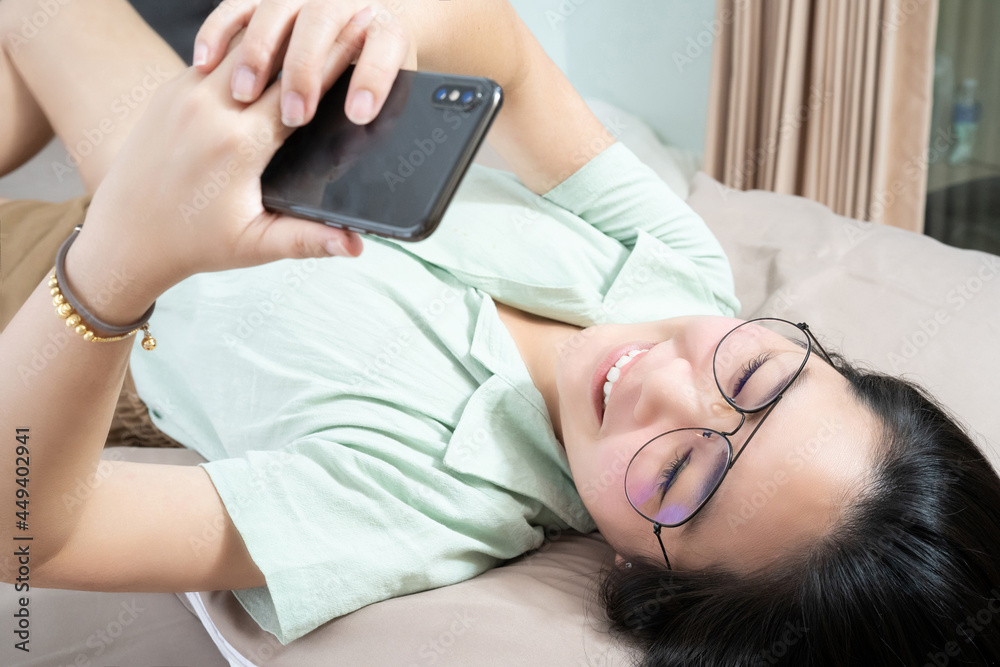 © Surachetsh - Beautiful glasses Asian woman lie on her back, chatting, typing, and texting, mobile in her bedroom with a pastel green - brown color theme. © Surachetsh - Beautiful glasses Asian woman lie on her back, chatting, typing, and texting, mobile in her bedroom with a pastel green - brown color theme.