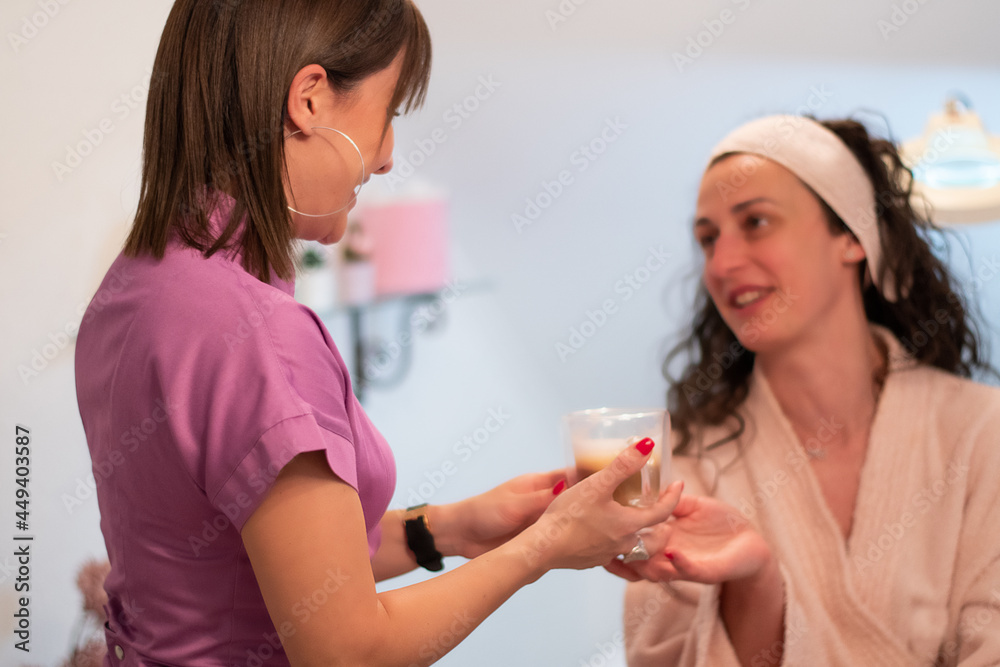 Attractive young beautician in purple uniform is serving cup of coffee  to her client after body treatment in beauty salon. Relaxing with beverages on cosmetic bed. Beauty , pleasure concept