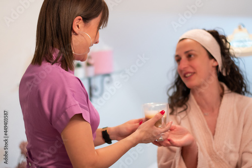 Attractive young beautician in purple uniform is serving cup of coffee to her client after body treatment in beauty salon. Relaxing with beverages on cosmetic bed. Beauty , pleasure concept
