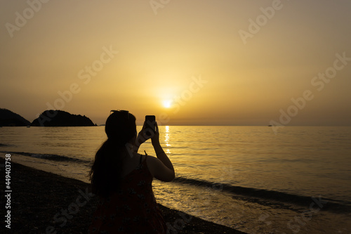 Silhouette of a person on the beach taking photograph with her mobile phone at dawn. Sun rises over the Mediterranean Sea on a lovely summer morning. Selective focus.